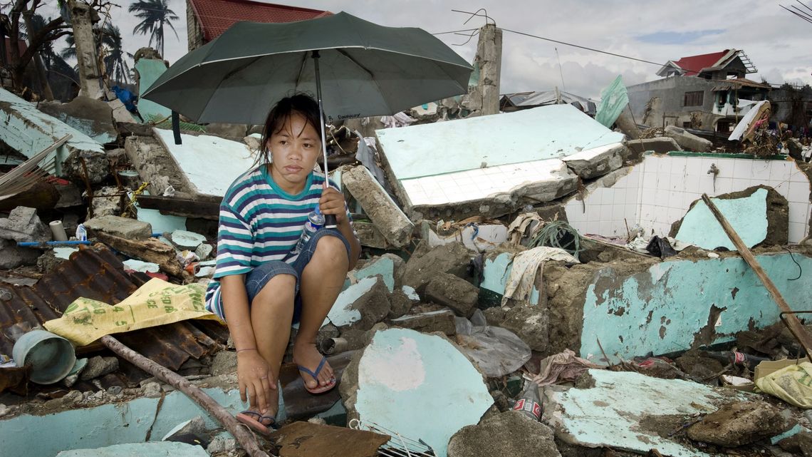 Frau mit einem Regenschirm hockt auf Trümmern ihres zerstörten Hauses